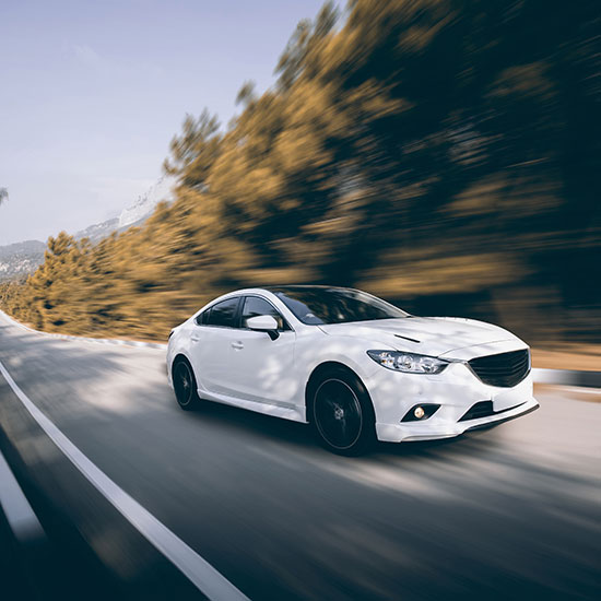 California’s Premier Dealerships Regain Momentum Post‑CARB Clarity; A white car speeds down a tree-lined road, creating a sense of motion. The blurred background of trees and mountains suggests fast travel.