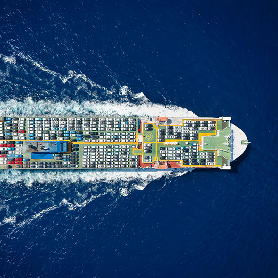 The Top 20 Trends in U.S. Auto Distribution for 2026; Aerial view of a large cargo ship moving through deep blue water, loaded with numerous vehicles. It creates white waves as it cuts through the ocean.